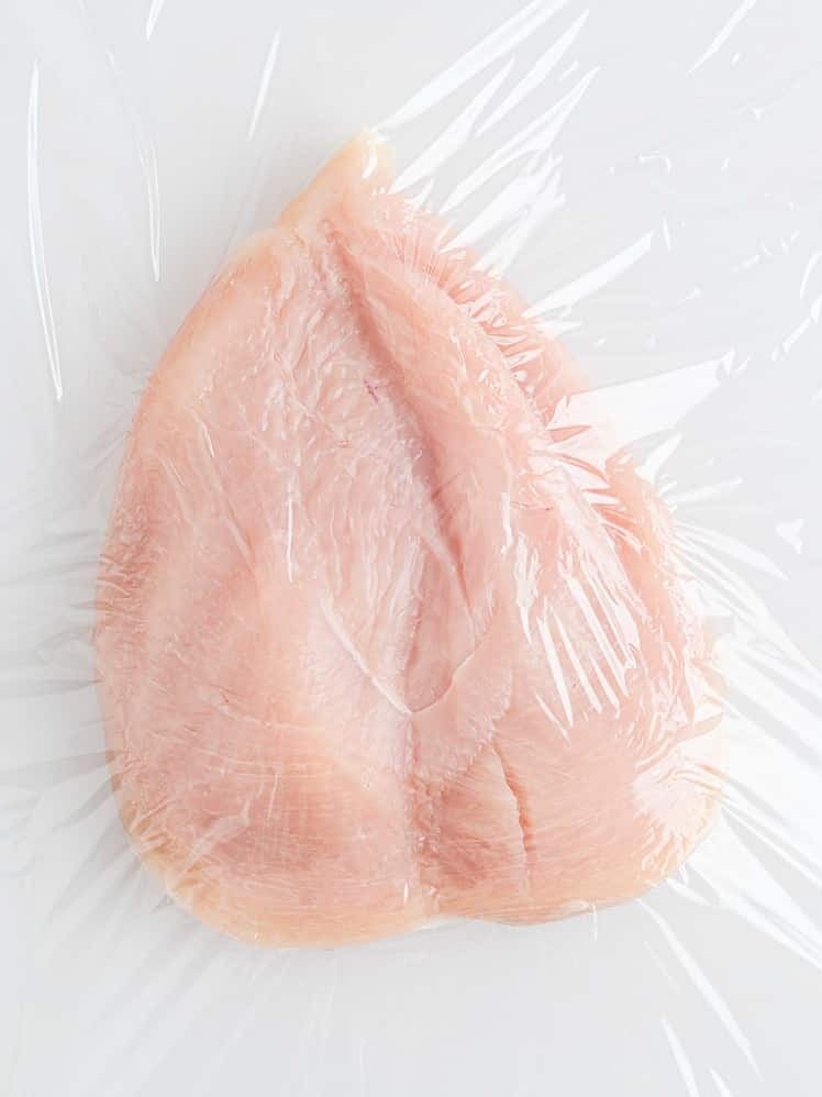 Raw boneless chicken breast covered with clear plastic wrap on a white surface.