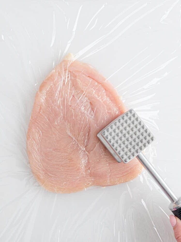 A raw chicken breast covered with plastic wrap is being tenderized with a metal meat mallet on a white surface.