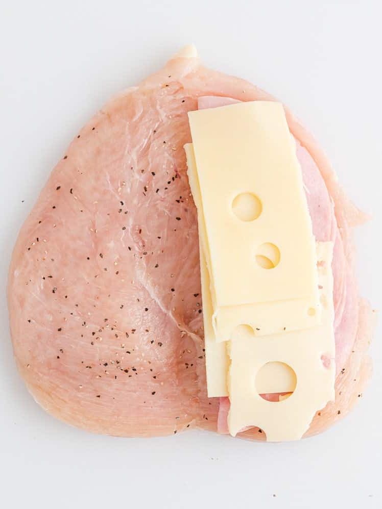 Raw chicken breast seasoned with pepper, topped with slices of ham and Swiss cheese, on a white background.