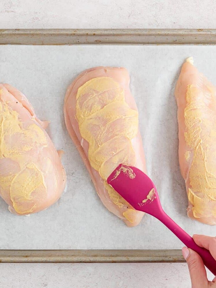 Three raw chicken breasts on a parchment-lined baking sheet; a hand spreads a yellow marinade or sauce on one breast with a pink spatula.