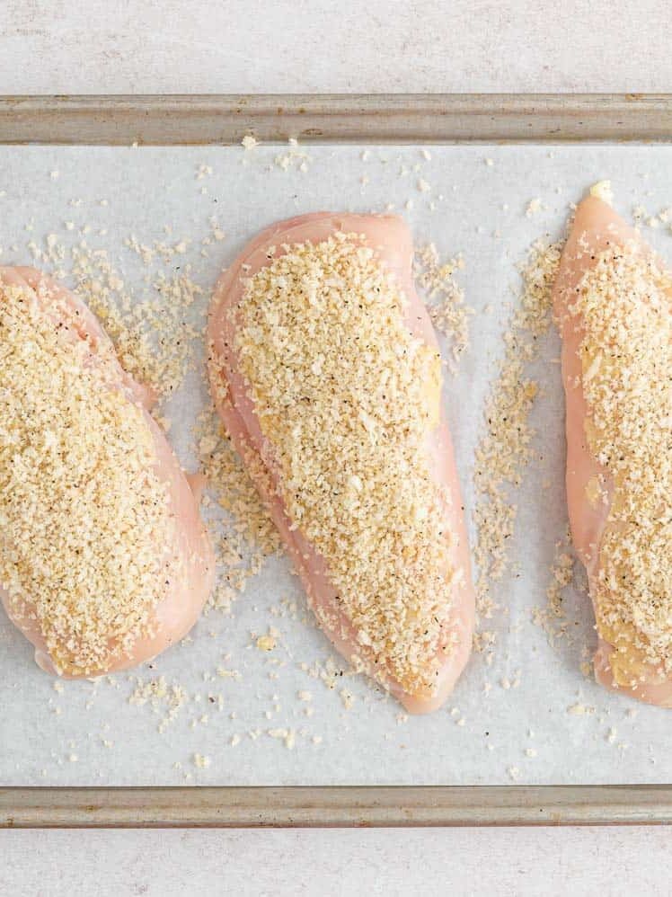 Three raw chicken breasts coated with breadcrumbs are arranged on a parchment-lined baking sheet.