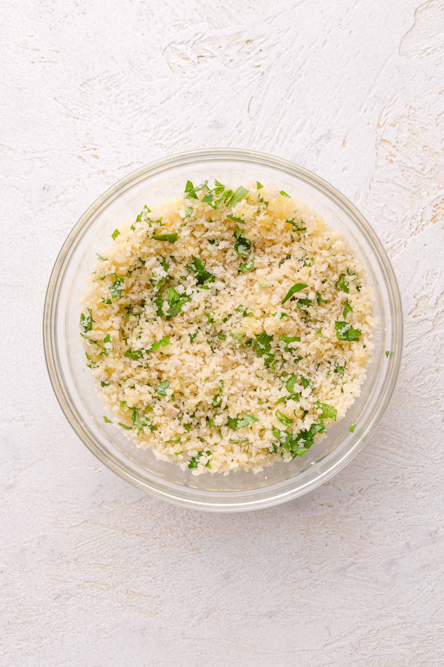 A clear glass bowl filled with a mixture of breadcrumbs and chopped parsley on a light textured surface.