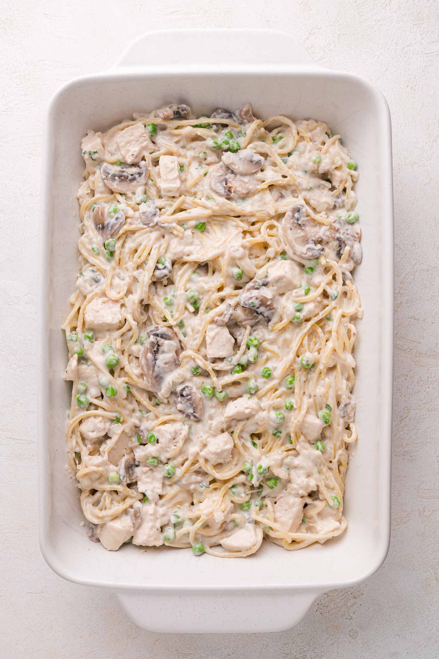 A white baking dish filled with creamy spaghetti, chicken pieces, mushrooms, and green peas on a light background.