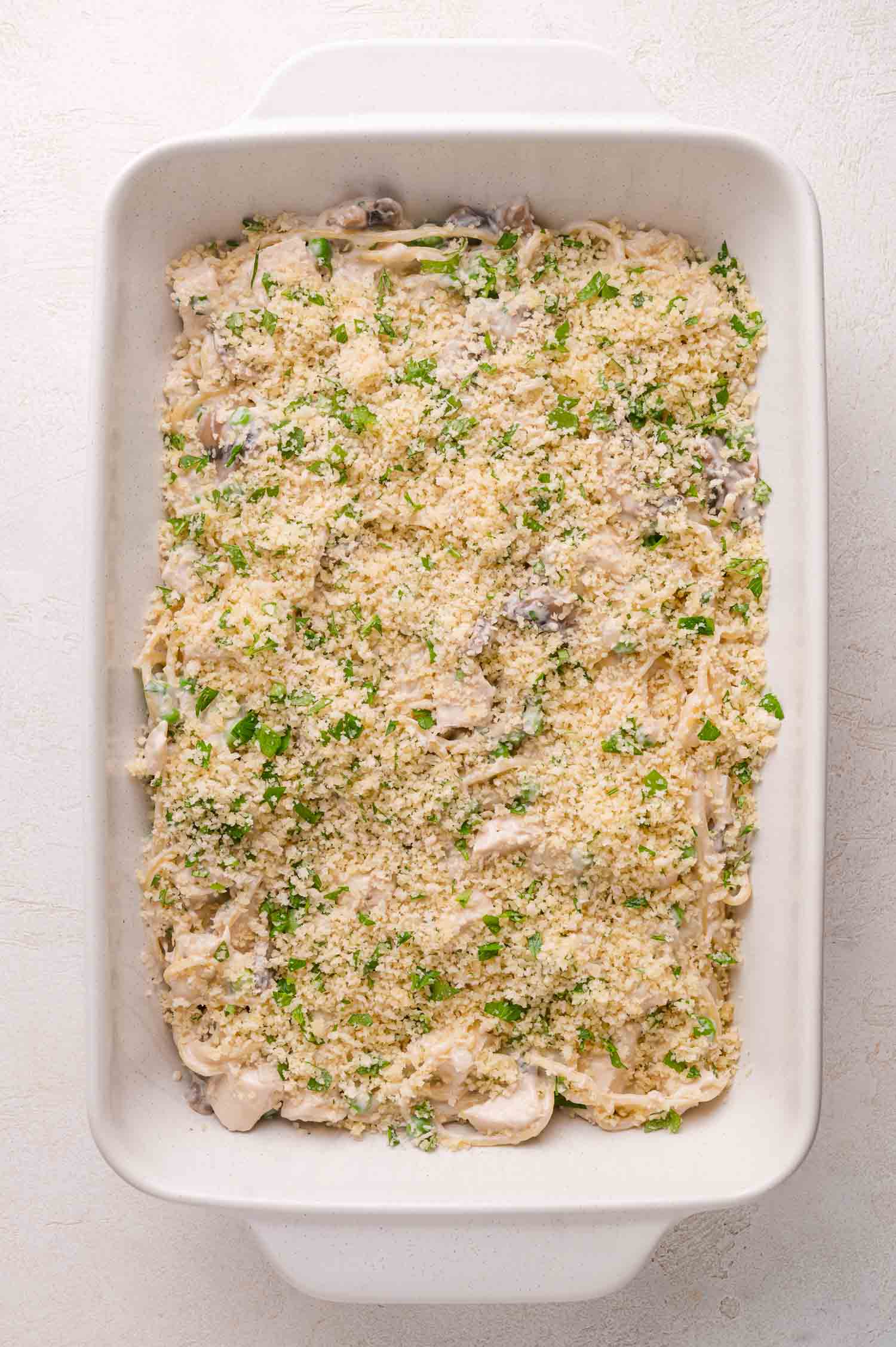 A white baking dish filled with an unbaked casserole topped with breadcrumbs and chopped herbs on a light surface.