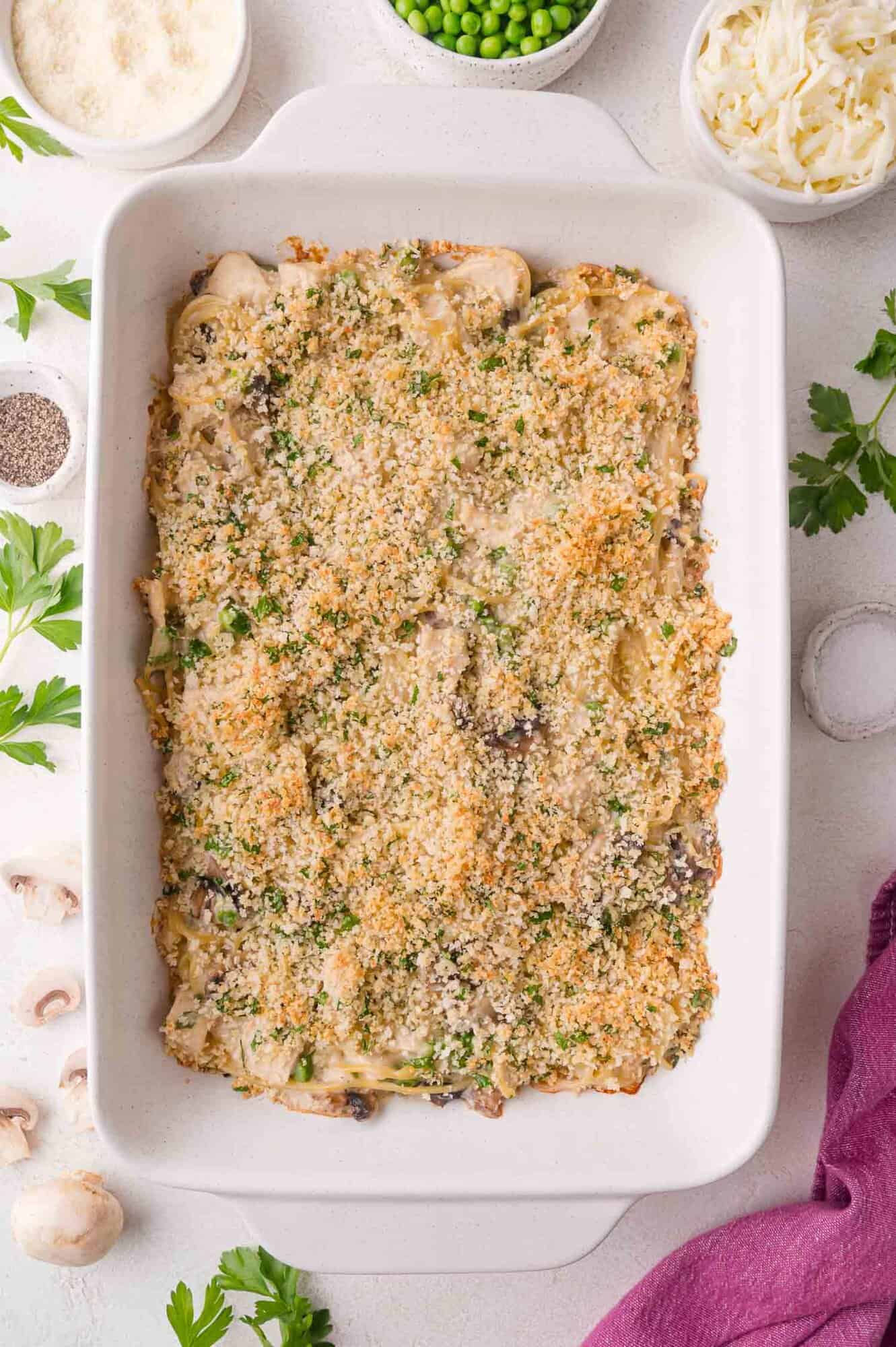 A baked chicken tetrazzini casserole topped with golden breadcrumbs sits in a white dish, surrounded by ingredients such as mushrooms, parsley, cheese, and peas.