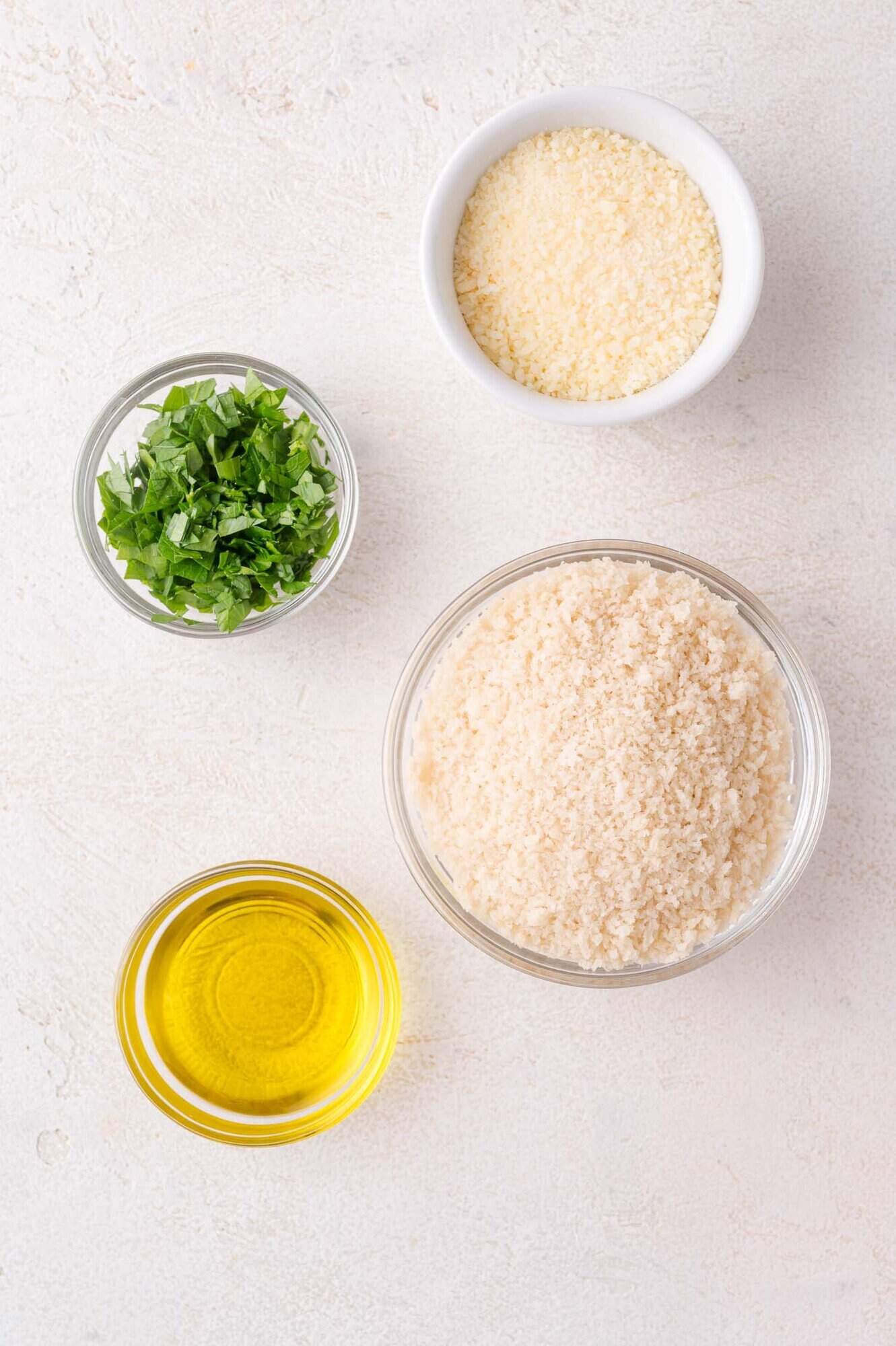 Four small bowls contain panko breadcrumbs, grated parmesan cheese, chopped parsley, and olive oil arranged on a light surface.