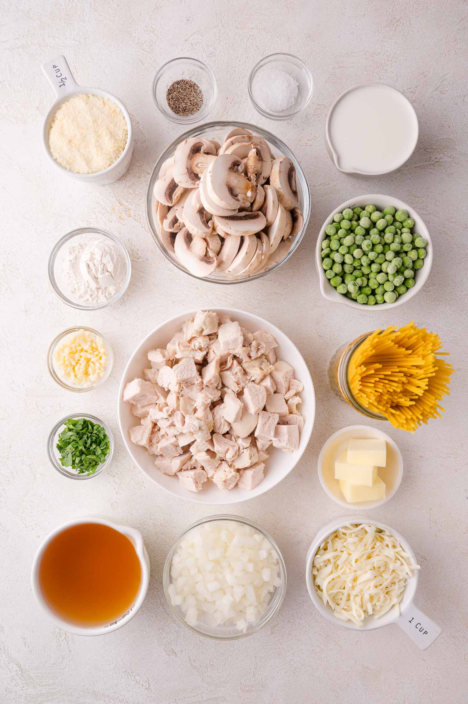 Overhead view of measured ingredients in bowls, including cooked chicken, mushrooms, peas, pasta, cheese, broth, butter, milk, onions, flour, garlic, and seasonings on a white surface.
