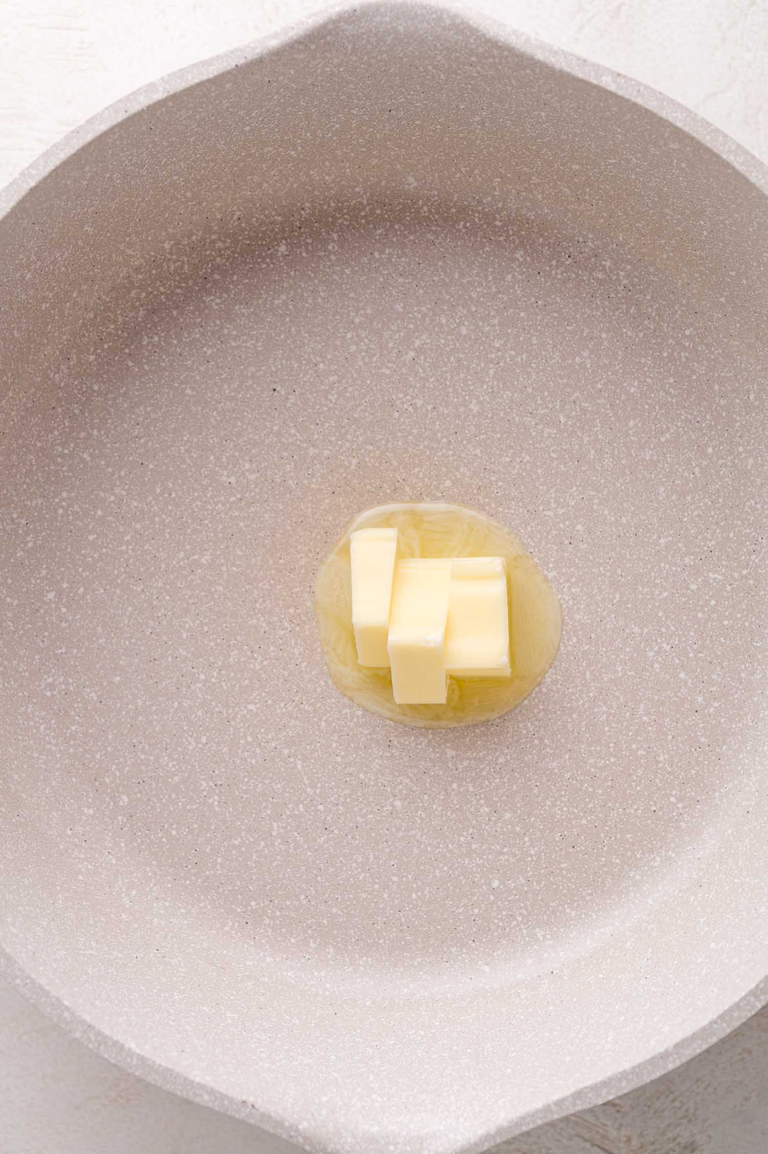 A few pieces of butter melting in the center of a light-colored, empty skillet.