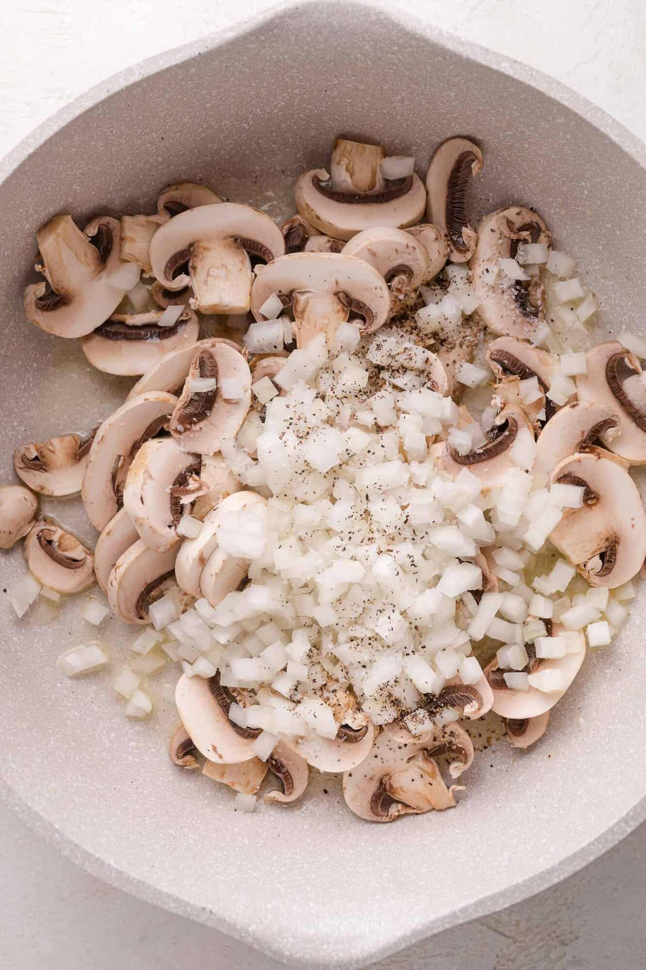 Sliced mushrooms and chopped onions seasoned with salt and pepper in a white frying pan.