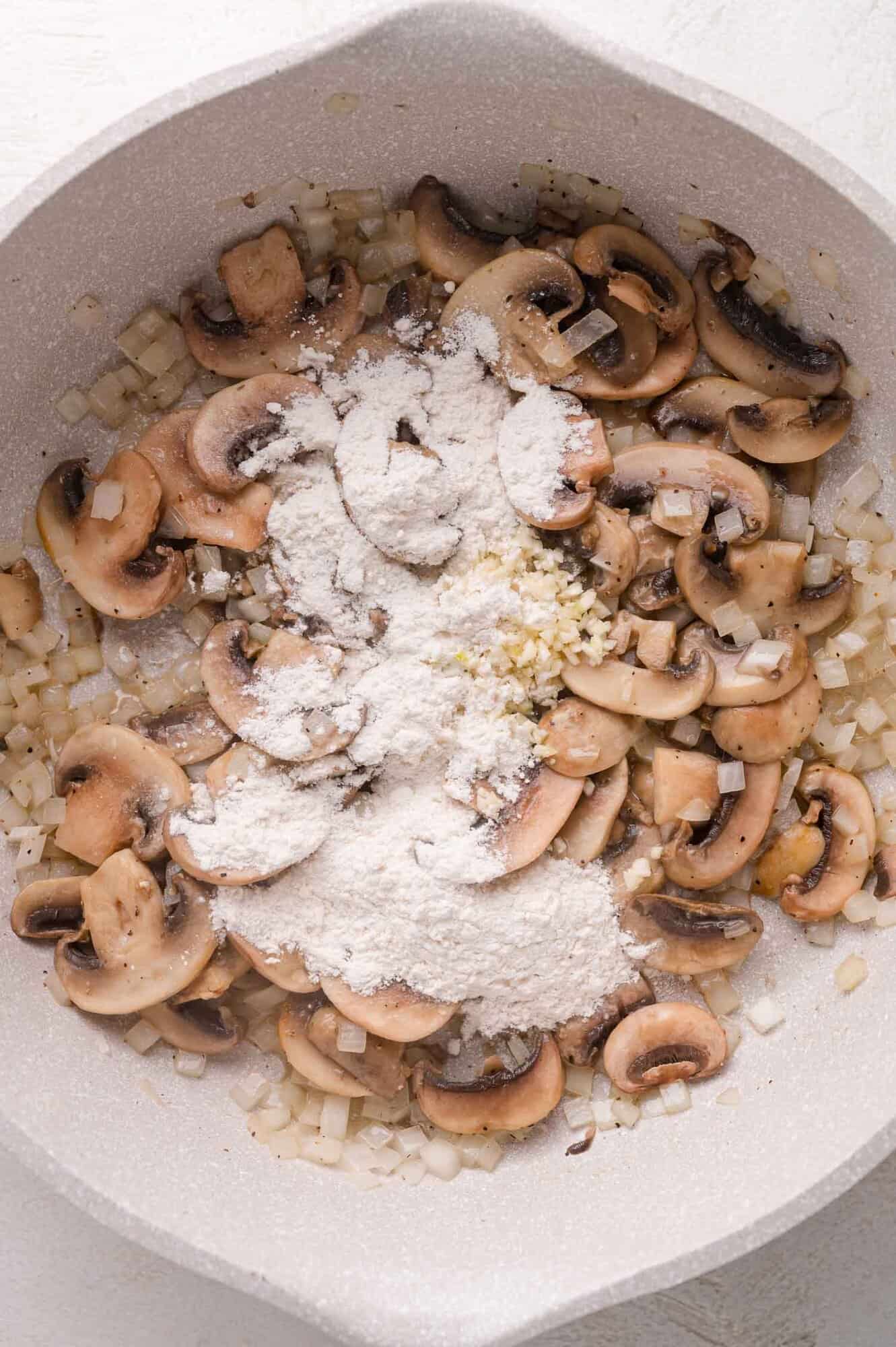 Sliced mushrooms, chopped onions, minced garlic, and flour cooking together in a light-colored skillet.