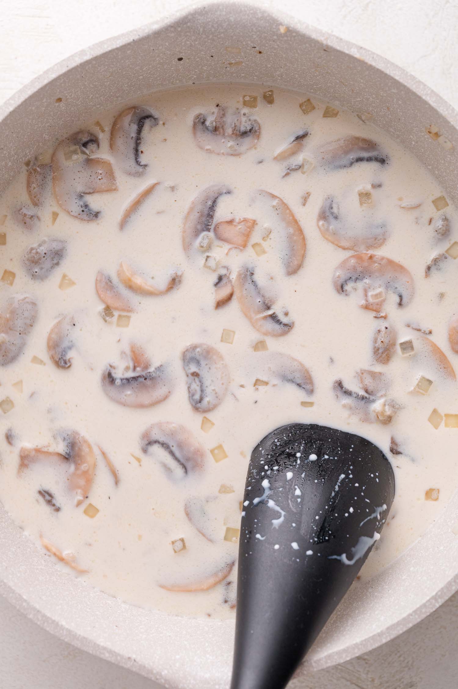 A creamy mushroom sauce with sliced mushrooms and diced onions is being stirred in a white pan with a black spoon.