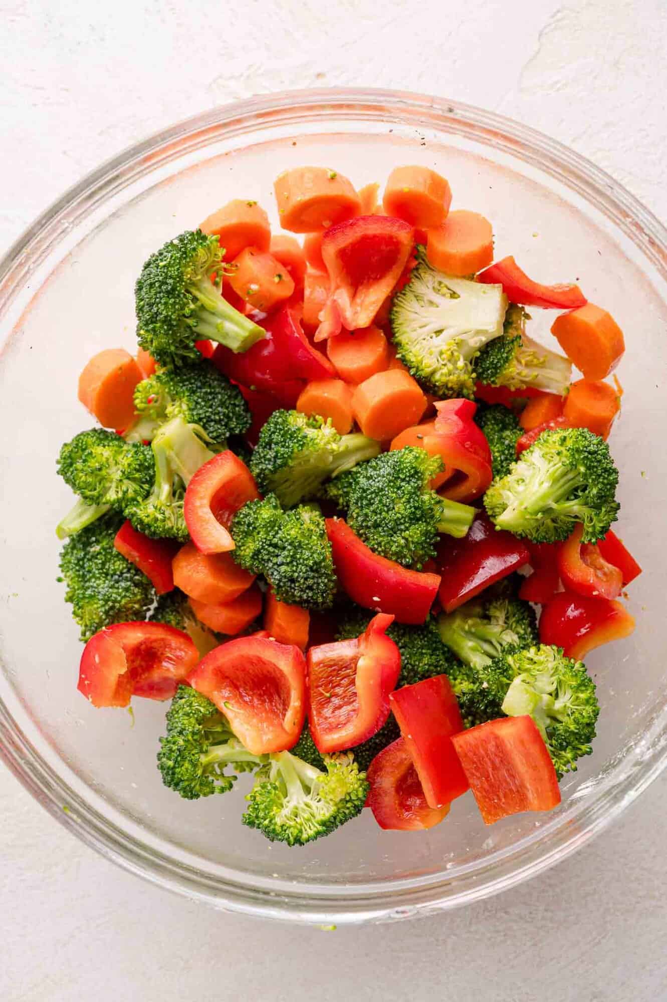 A glass bowl filled with chopped broccoli, carrots, and red bell peppers on a white surface.