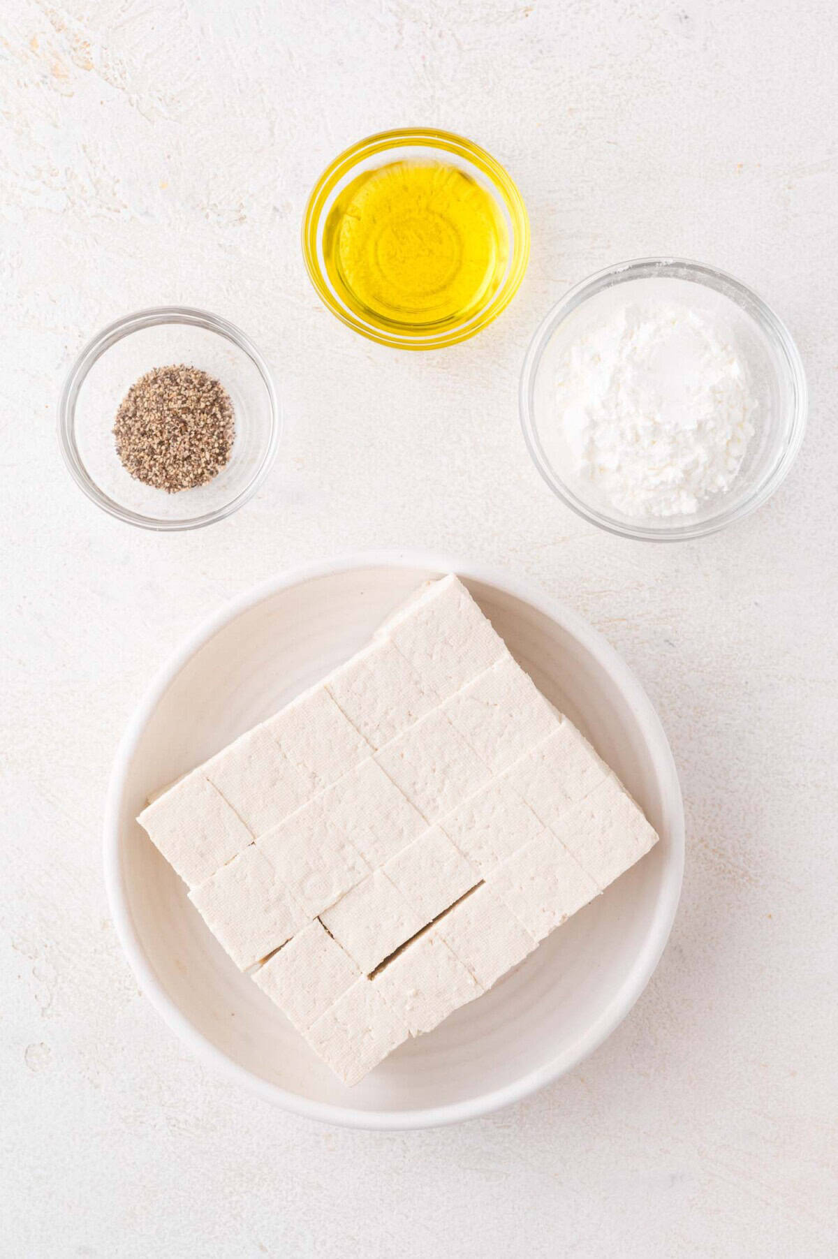 A bowl of cubed tofu, a small bowl of oil, a bowl of cornstarch, and a bowl of ground black pepper arranged on a white surface.