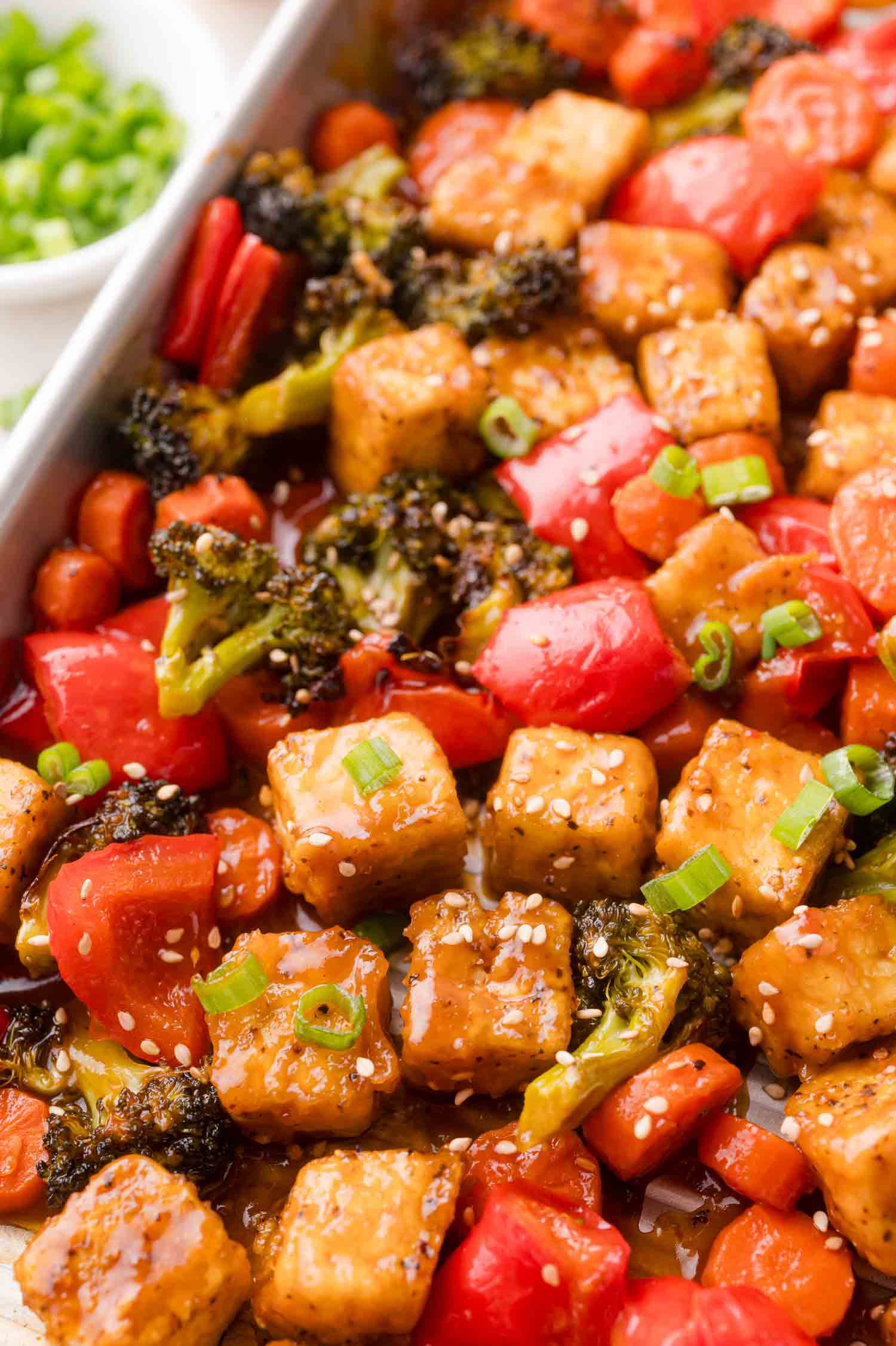 A close-up of a tray filled with baked tofu cubes, broccoli, red bell pepper, and carrots, garnished with sesame seeds and chopped green onions.