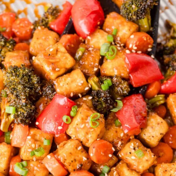 Sheet pan General Tso tofu with broccoli, red bell peppers, and carrots, garnished with sesame seeds and chopped green onions.