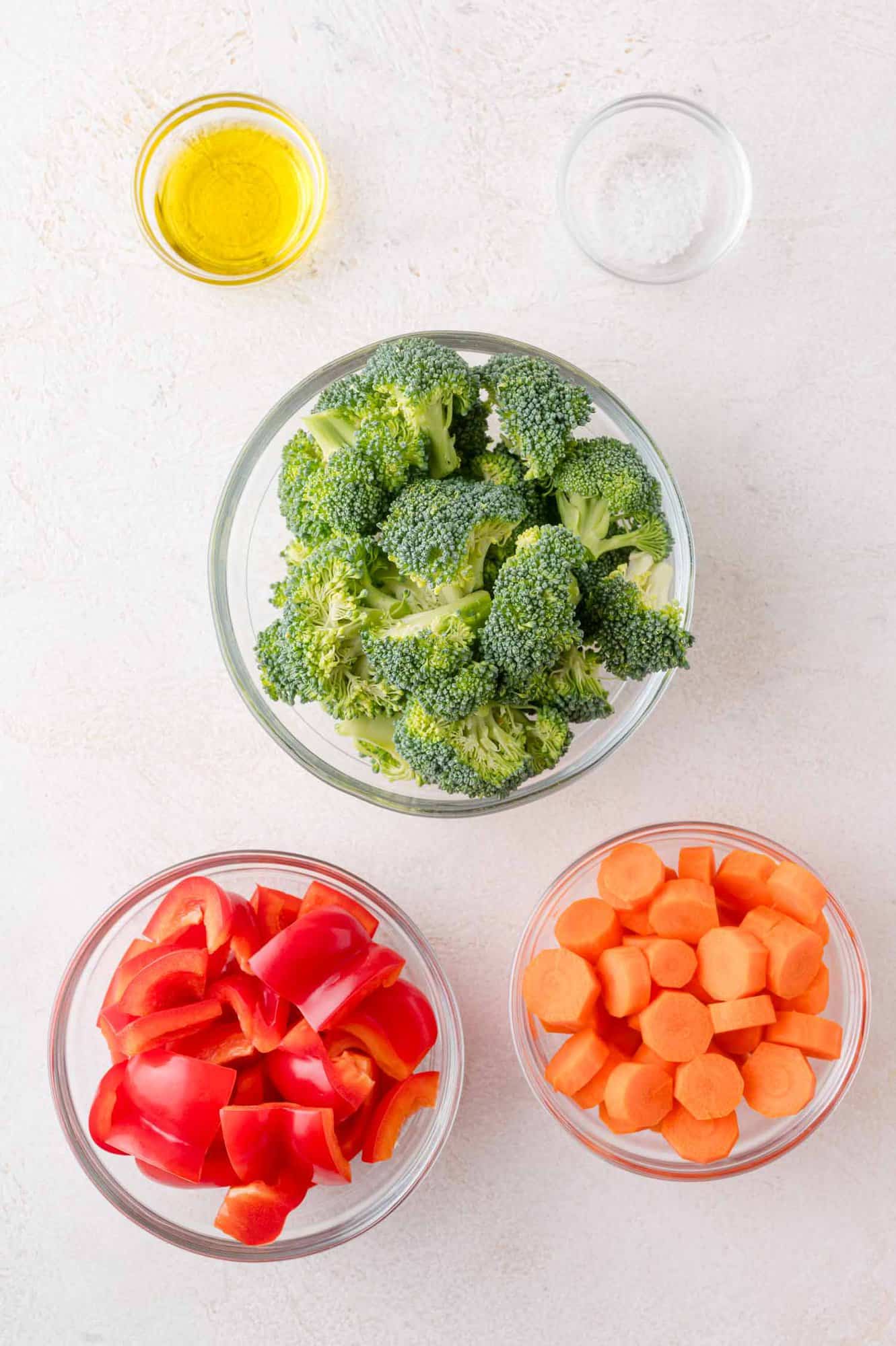 Three bowls containing broccoli florets, chopped red bell peppers, and sliced carrots, with small bowls of olive oil and salt on a white surface.