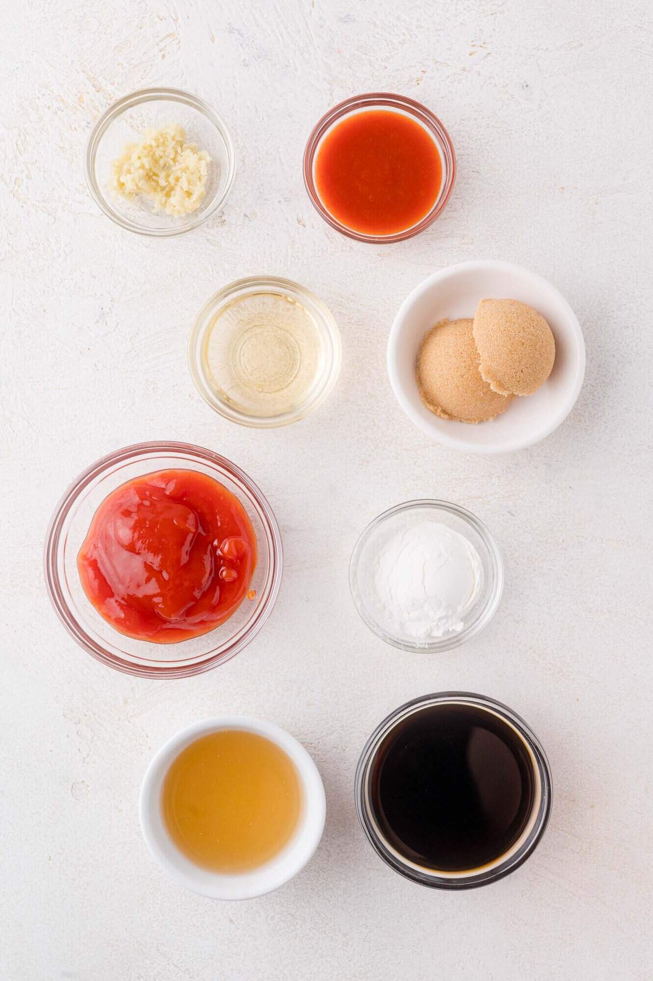 Eight small bowls containing various ingredients, including ketchup, brown sugar, soy sauce, honey, minced garlic, vinegar, cornstarch, and a red sauce, arranged on a white surface.