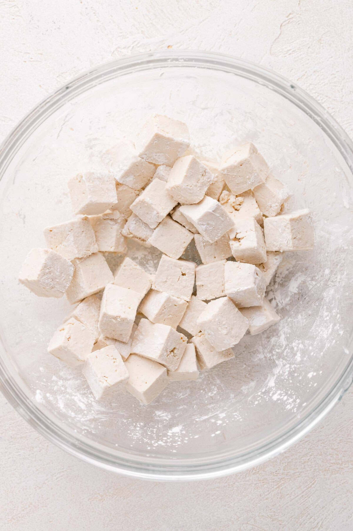 Glass bowl containing cubed pieces of tofu, lightly coated with cornstarch, on a light-colored surface.