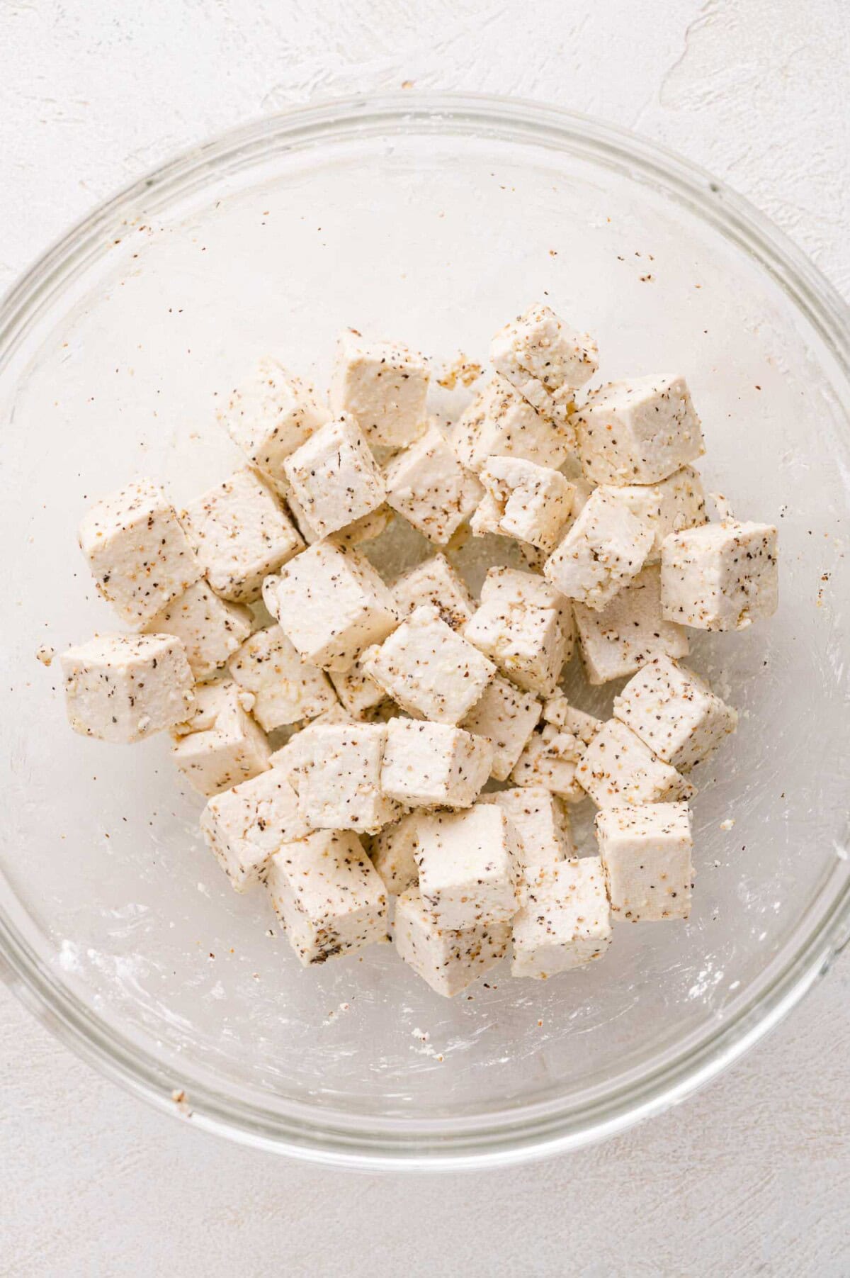 A glass bowl containing cubed tofu coated with black pepper and seasoning on a white surface.