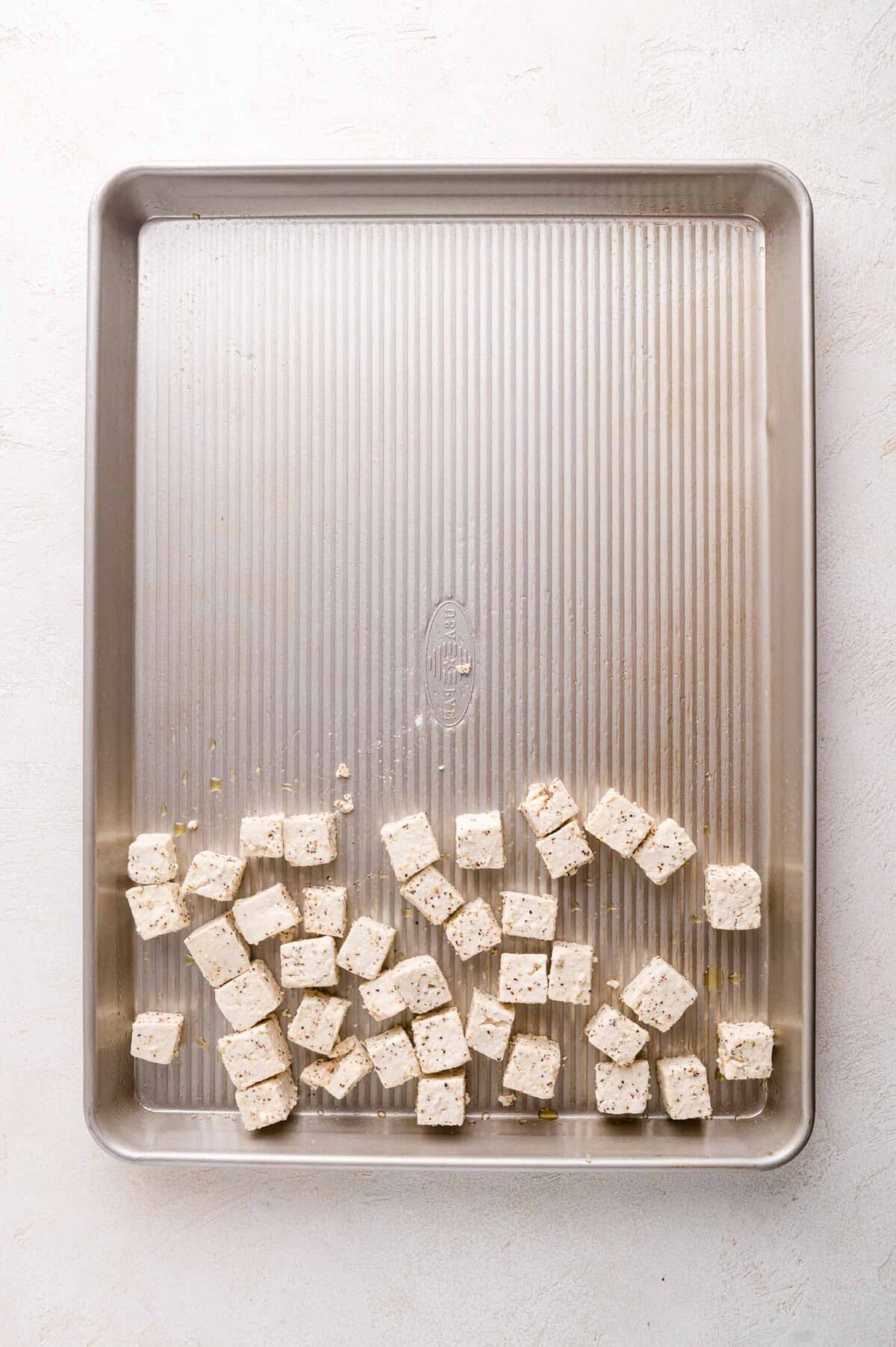 Cubed tofu scattered on one side of a large, empty metal baking sheet on a light-colored surface.