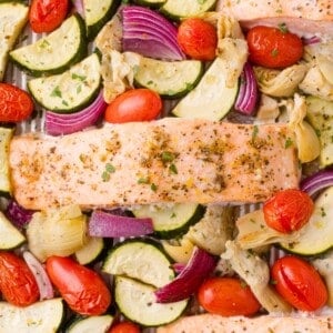 Baked salmon fillets on a sheet pan with zucchini, red onion, cherry tomatoes, and artichoke hearts, seasoned with herbs.