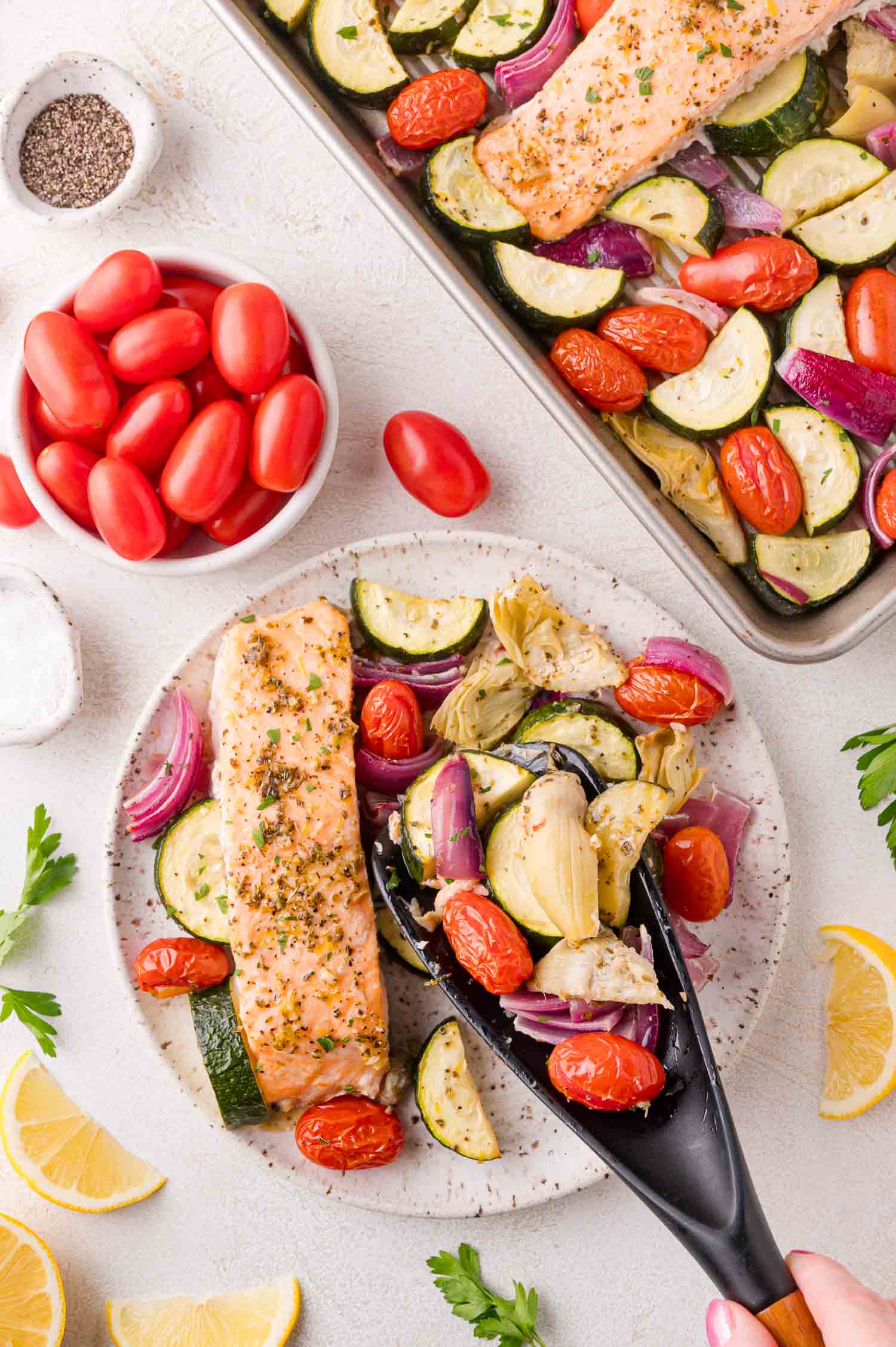 A plate with baked salmon, zucchini, tomatoes, red onion, and artichokes is served; a tray of the same dish and a bowl of grape tomatoes are nearby.