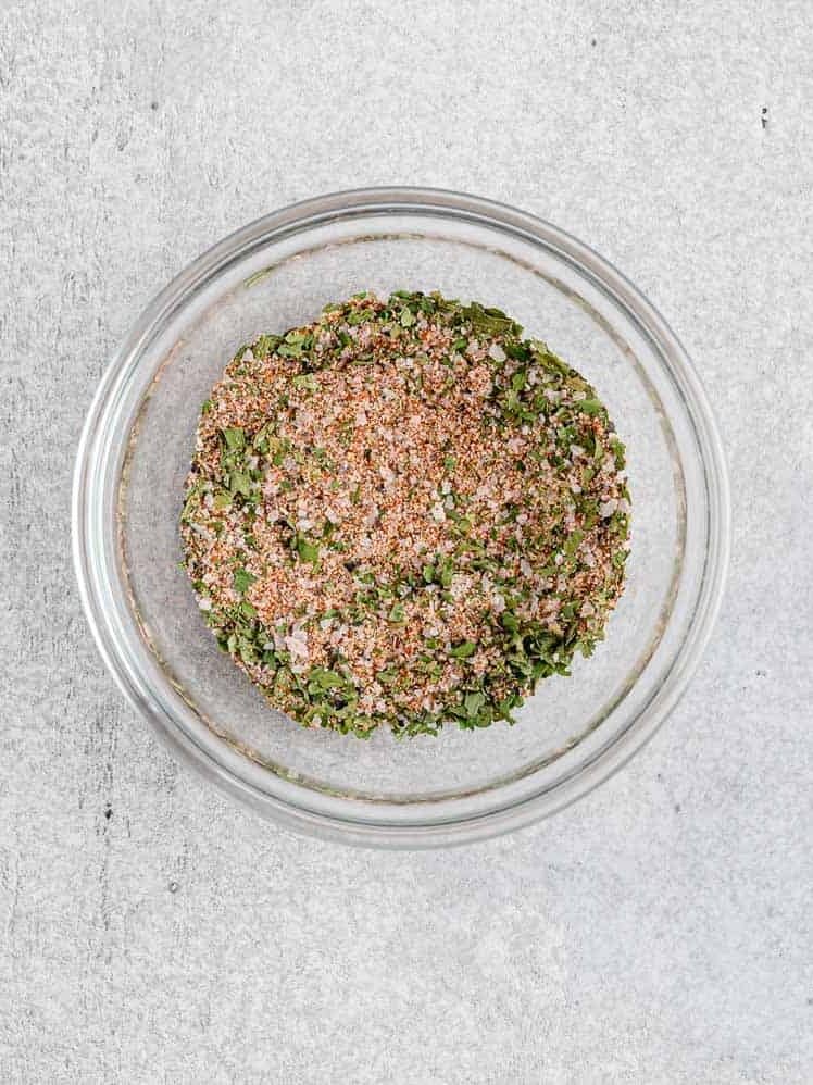 A glass bowl filled with a mixture of dried herbs and spices on a light gray surface.