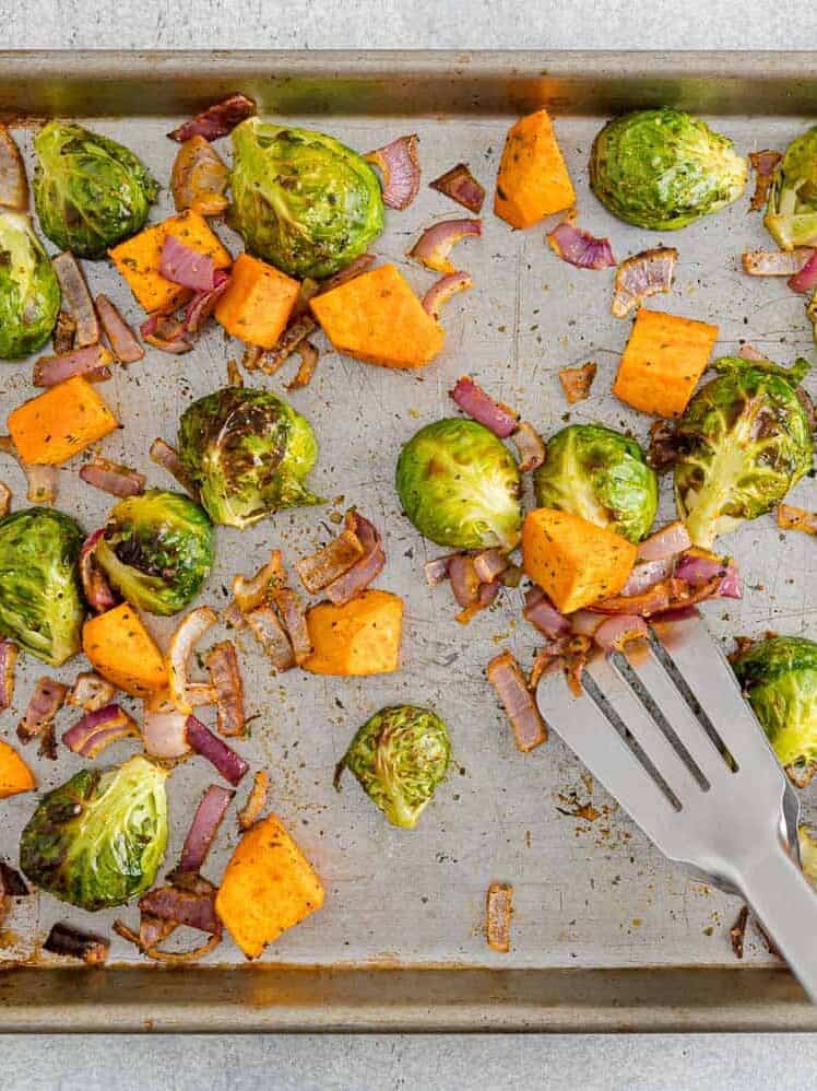 Roasted Brussels sprouts, sweet potato cubes, and red onion pieces are scattered on a baking sheet with a metal spatula.