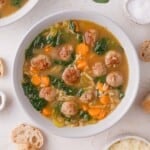 A bowl of soup containing meatballs, orzo pasta, spinach, sliced carrots, and celery in a clear broth, with pieces of bread on the side.