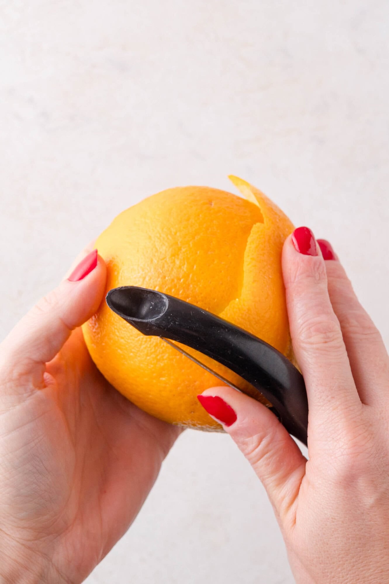 A person with red-painted nails uses a black peeler to remove the peel from an orange.