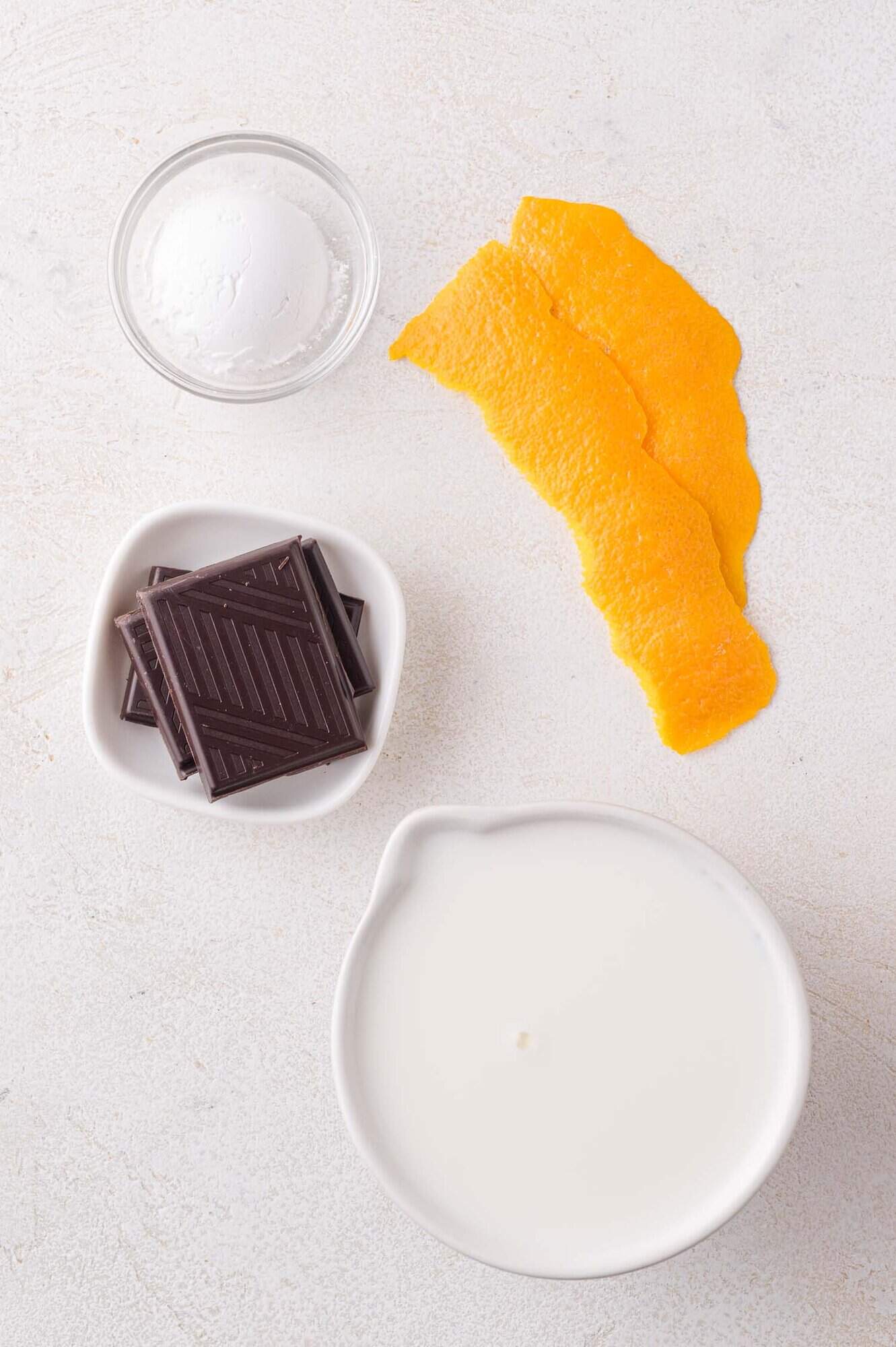 A bowl of cream, chocolate squares, orange peels, and a small bowl of powdered sugar are arranged on a white surface.