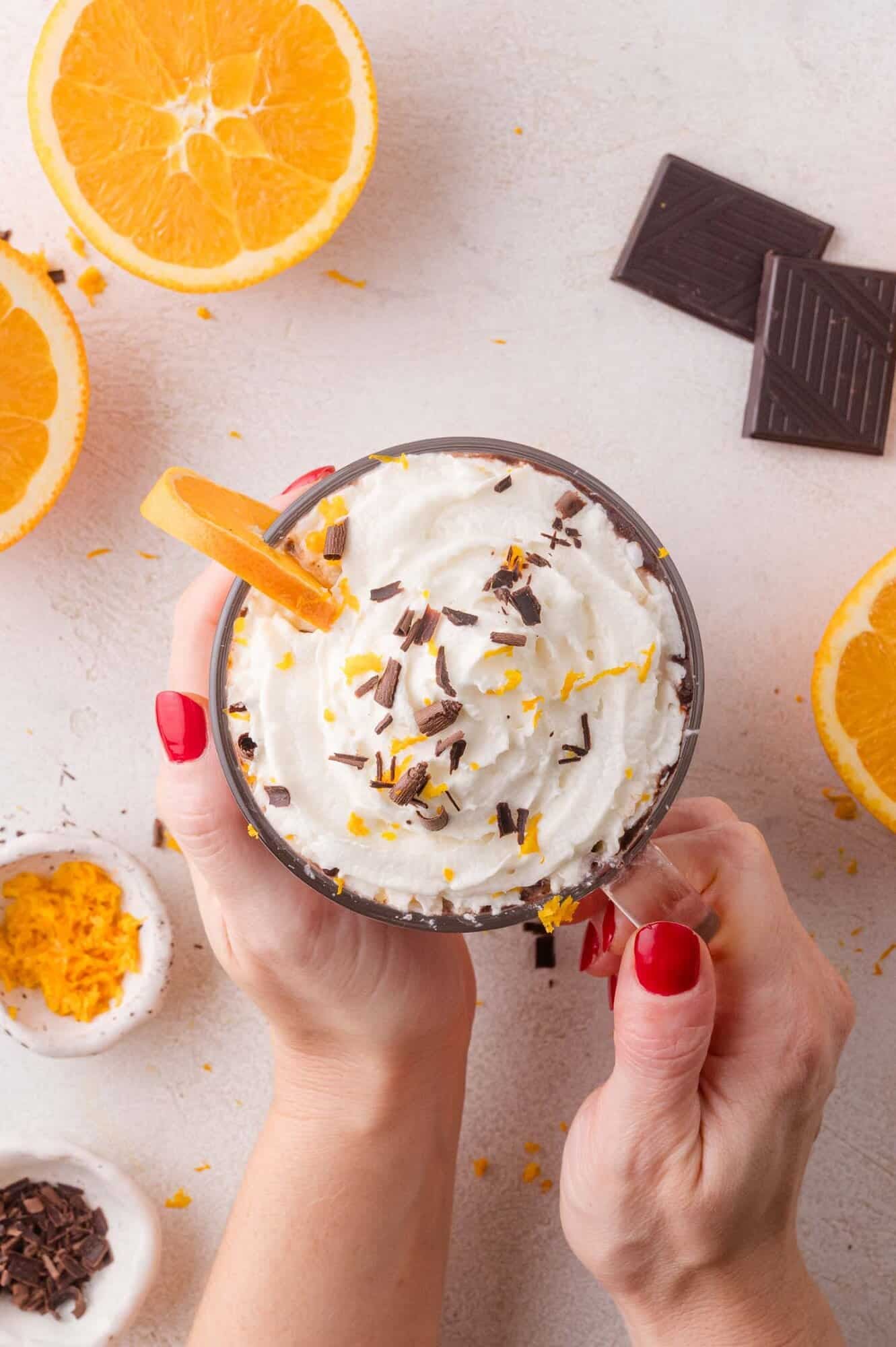 A person with red nail polish holds a glass of hot chocolate with whipped cream topped with chocolate shavings and orange zest, surrounded by orange slices and chocolate pieces.