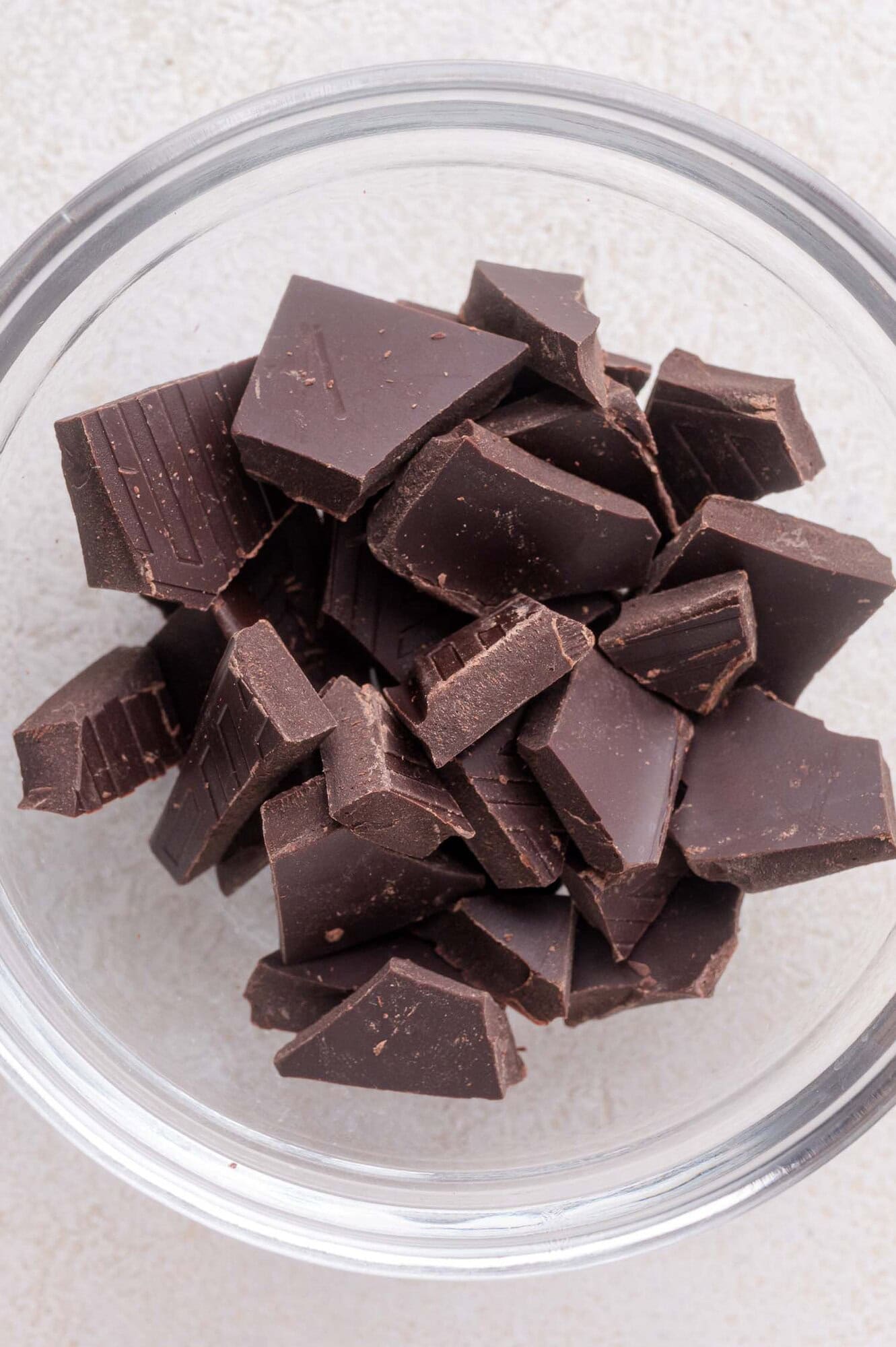 A clear glass bowl filled with irregularly chopped pieces of dark chocolate on a light surface.