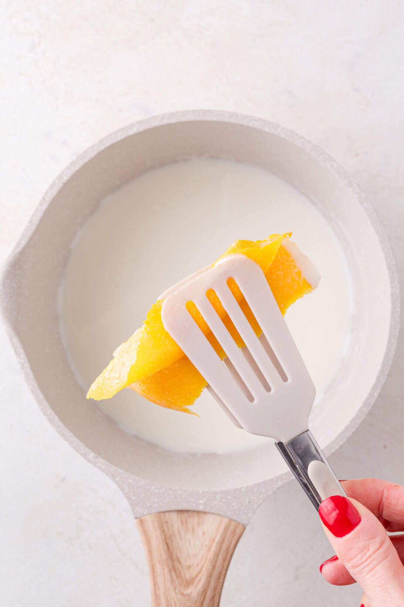 A hand holding tongs with orange peel over a saucepan containing milk.