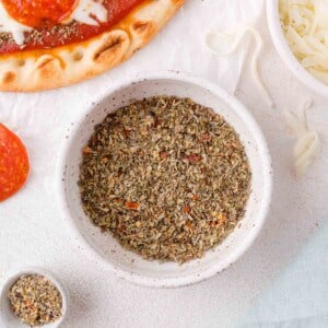 A bowl of dried Italian seasoning blend sits on a white surface next to pizza crust, shredded cheese, and a slice of pepperoni.