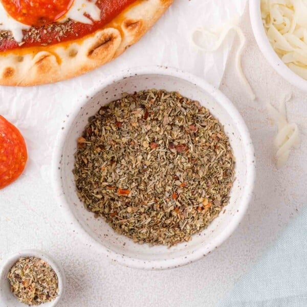 A bowl of dried Italian seasoning blend sits on a white surface next to pizza crust, shredded cheese, and a slice of pepperoni.