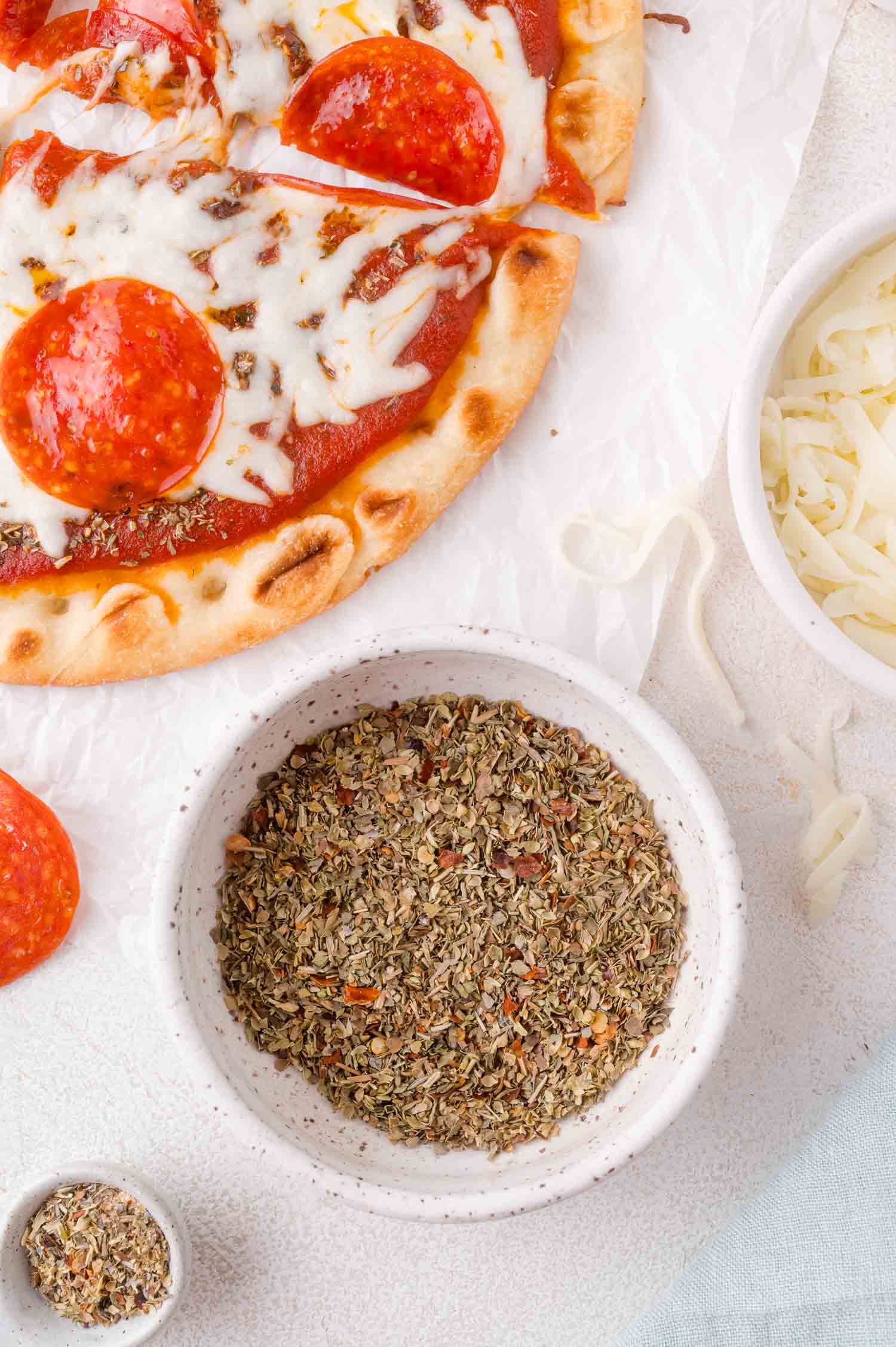 A pizza topped with melted cheese and pepperoni is next to a bowl of pizza seasoning and a bowl of shredded cheese on a white surface.