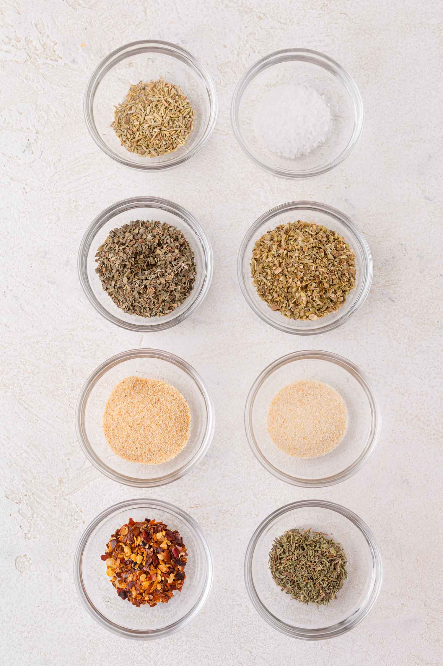 Eight small glass bowls filled with different dried herbs and spices, arranged in two vertical columns on a light surface.