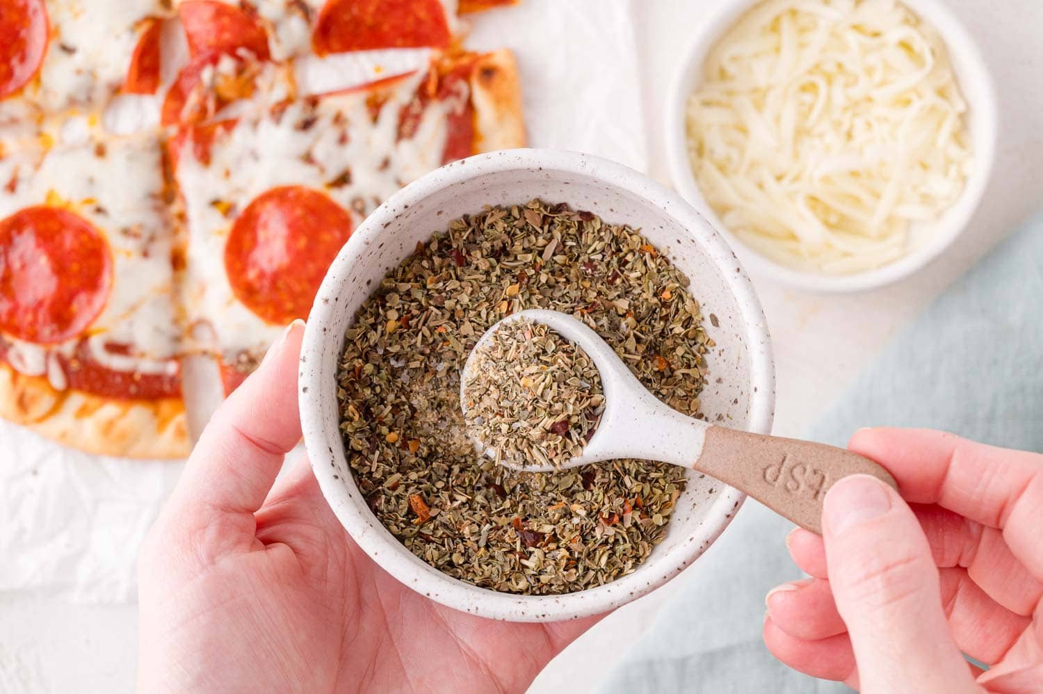 A hand holds a bowl of dried pizza seasoning with a measuring spoon, next to a pepperoni pizza and a small bowl of shredded cheese.