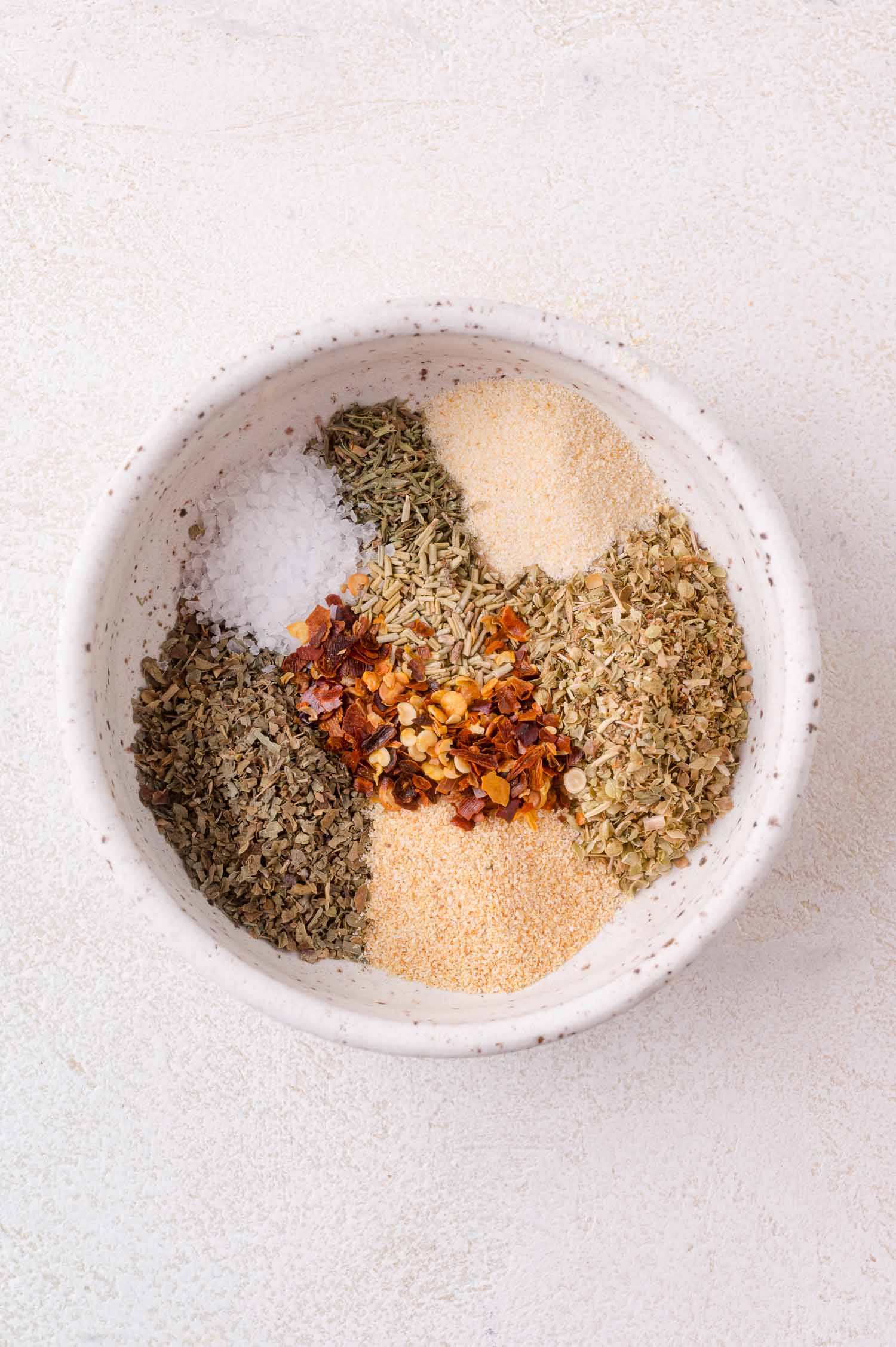 A white bowl containing separate piles of dried herbs, crushed red pepper flakes, salt, garlic powder, and onion powder on a light surface.