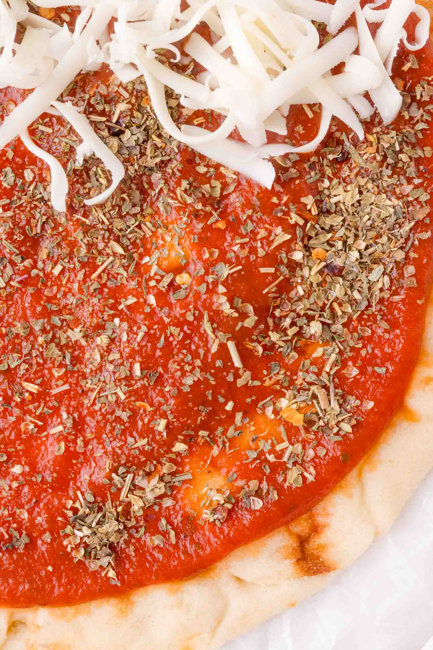 Close-up of pizza dough topped with tomato sauce, dried pizza seasoning, and shredded cheese on one side.