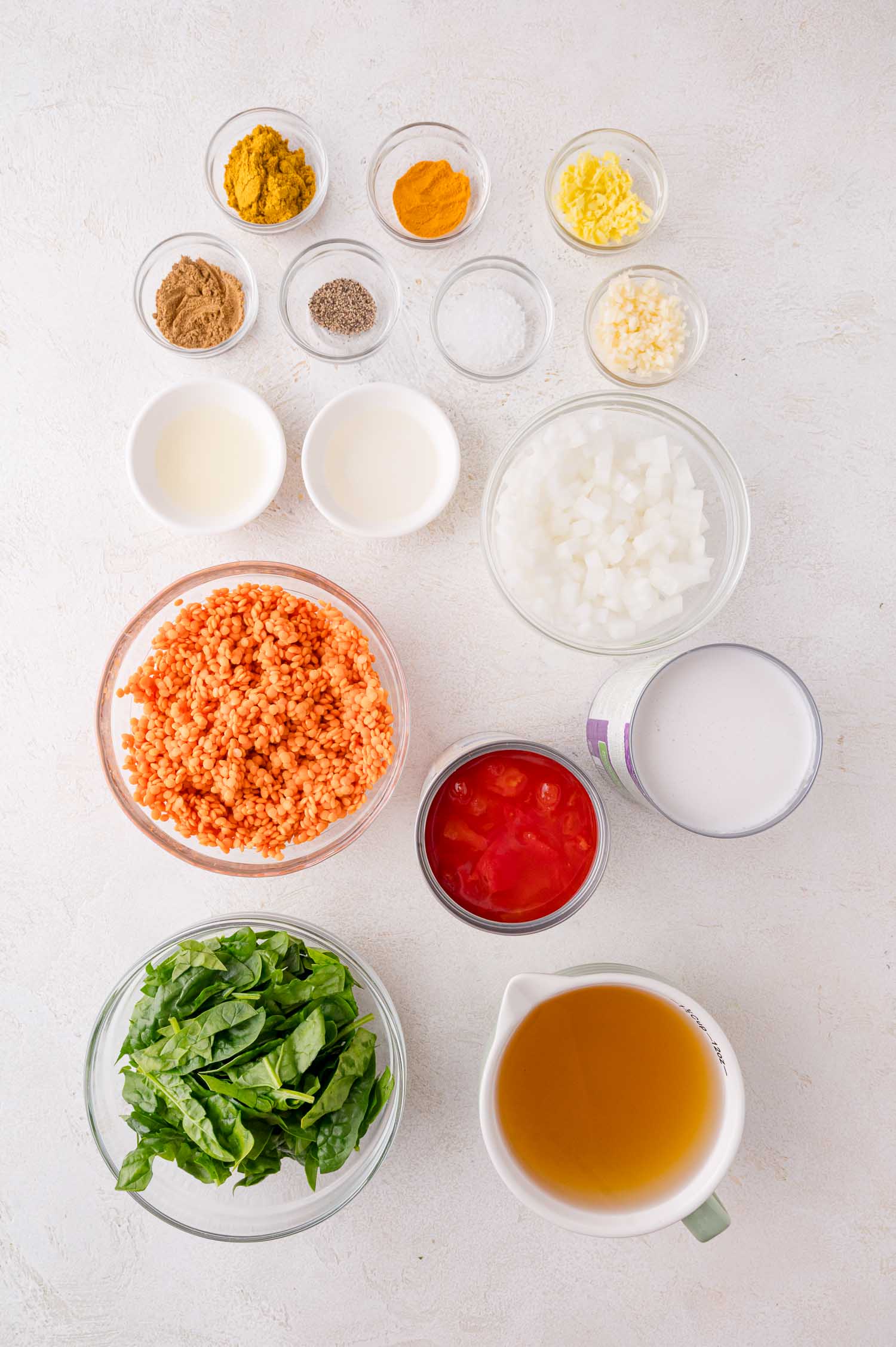 Ingredients for a lentil curry soup displayed in bowls and measuring cups, including spices, chopped onions, garlic, ginger, lentils, tomatoes, spinach, coconut milk, and broth.