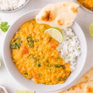 A bowl of white rice topped with red lentil curry, garnished with chopped herbs, a slice of lime, and a piece of naan bread.