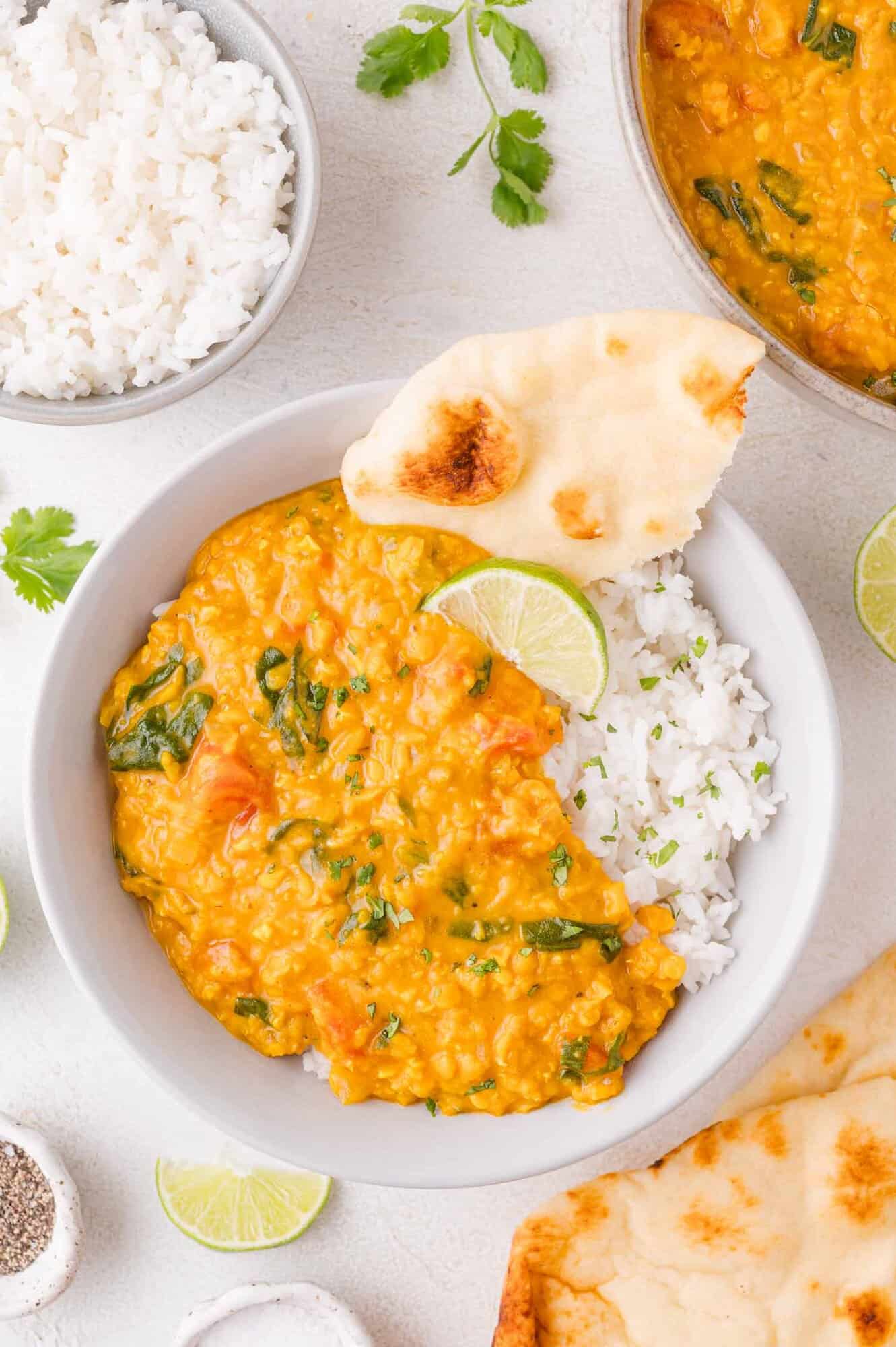 A bowl of white rice topped with red lentil curry, garnished with a lime wedge and served with naan bread. Other dishes of rice, curry, naan, and lime are nearby.