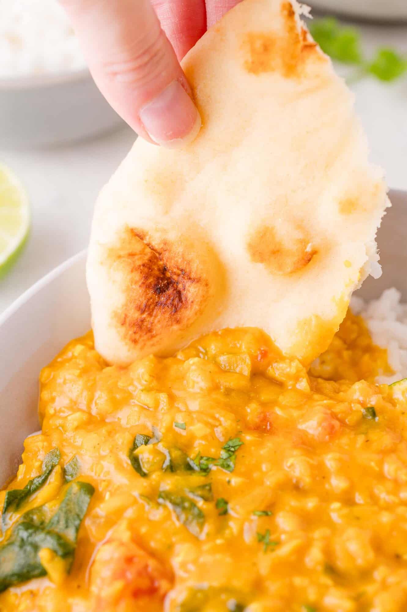 A hand holds a piece of naan bread, dipping it into a bowl of red lentil curry served over white rice.