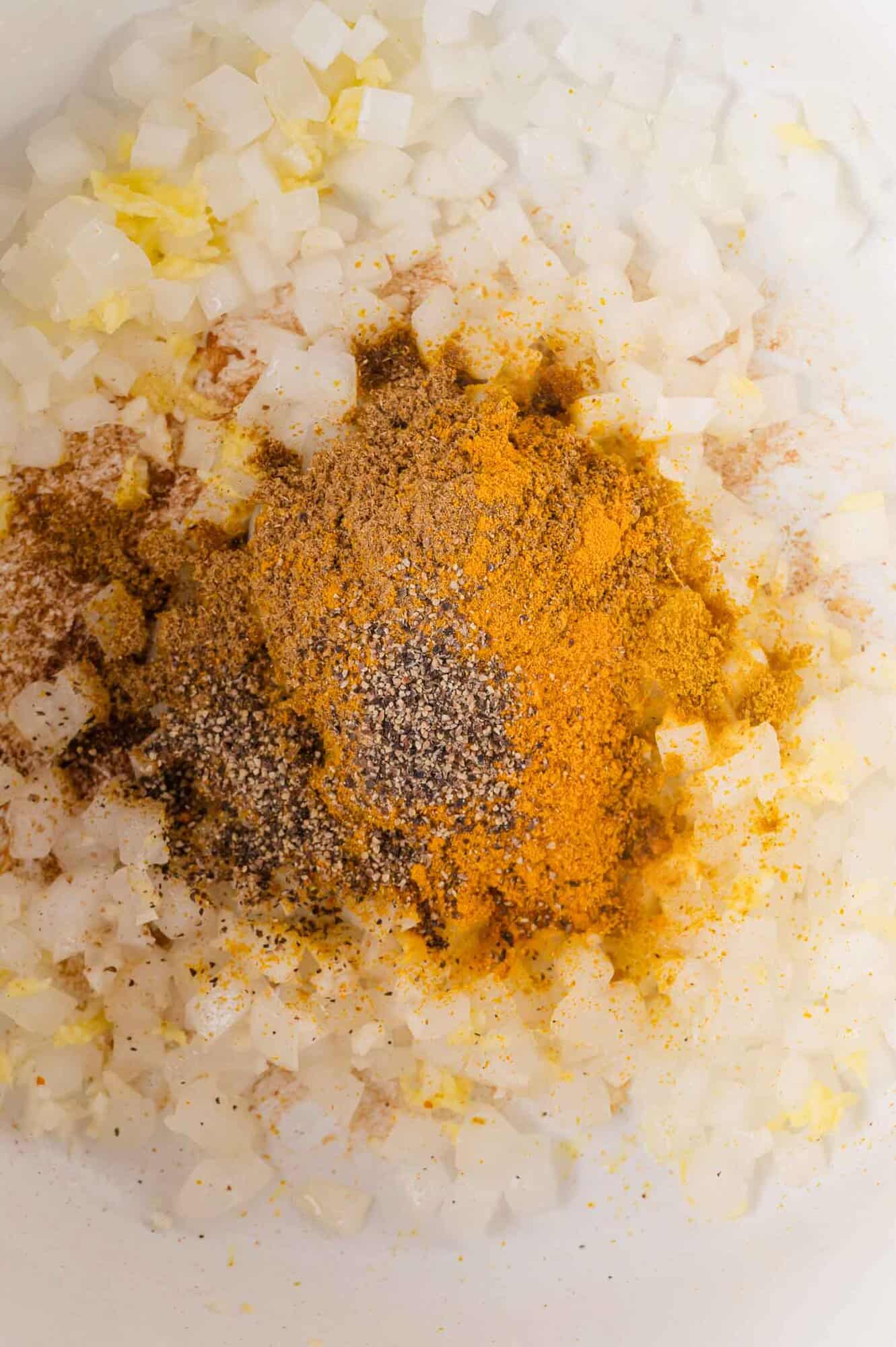 Chopped onions topped with ground black pepper, turmeric, and other spices in a white pan.