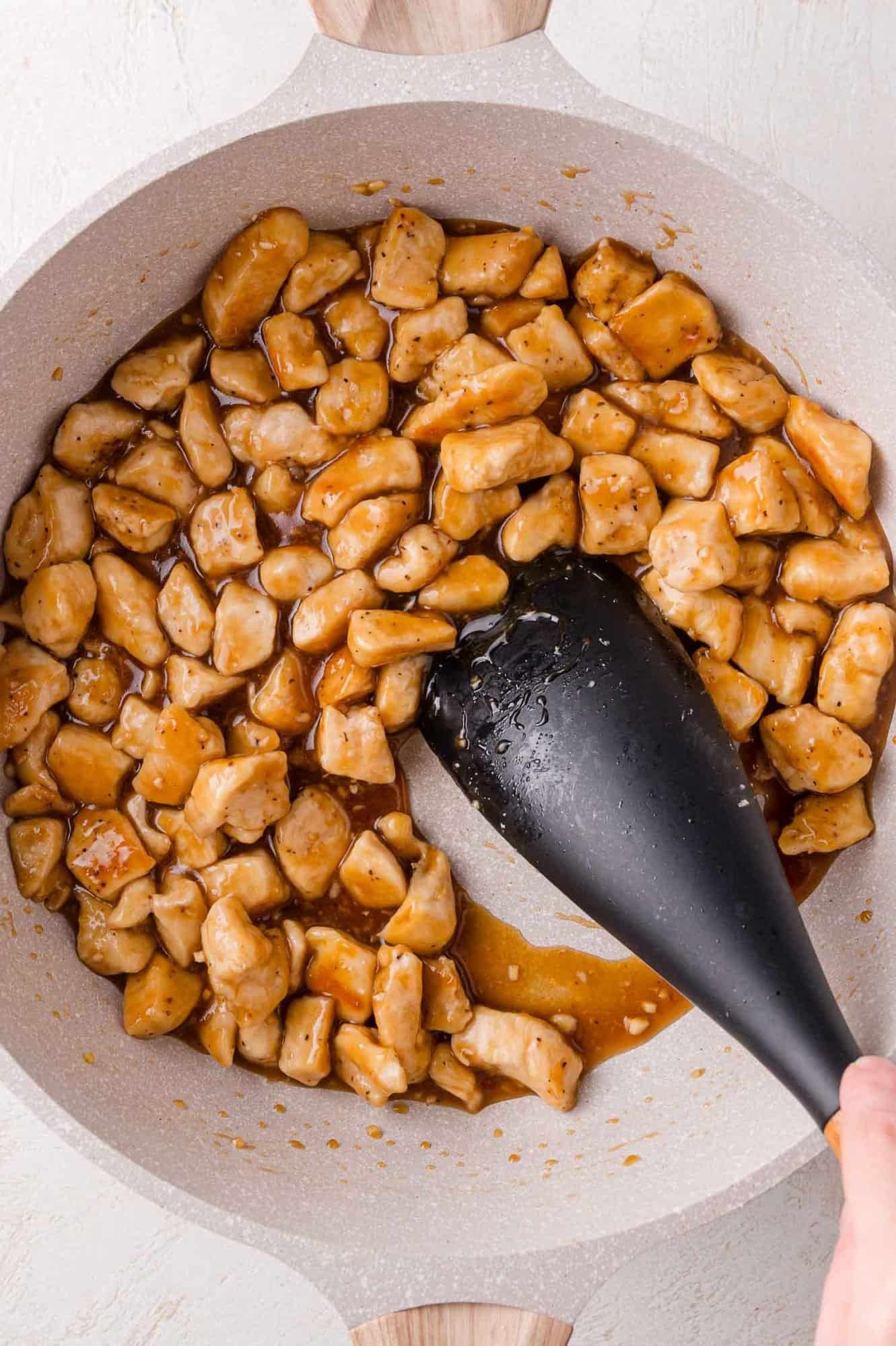 Chunks of chicken coated in brown teriyaki sauce being stirred with a black spatula in a light-colored pan.