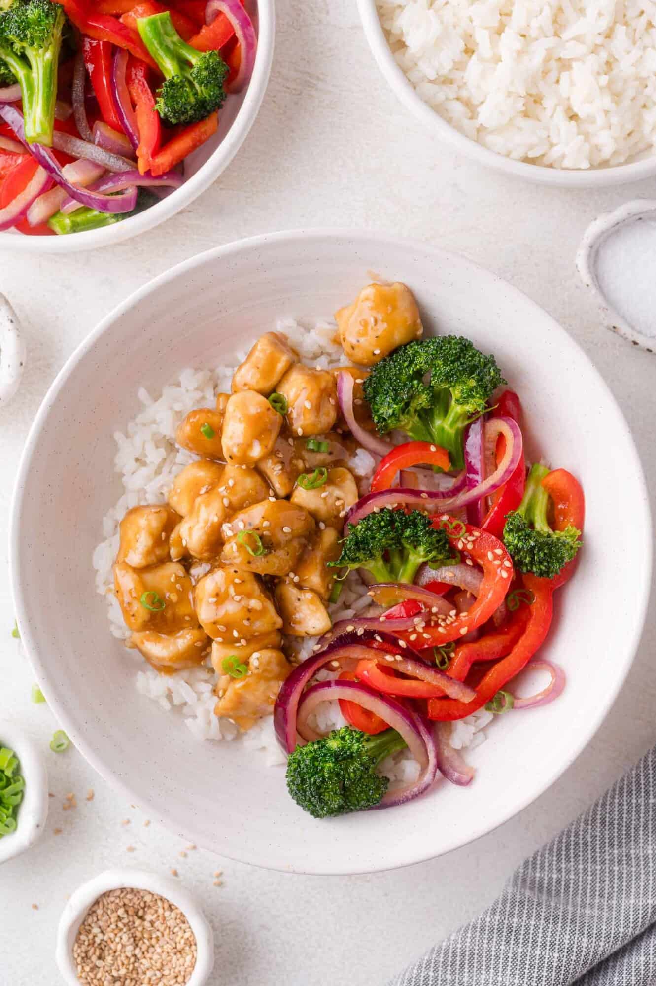 A bowl of white rice topped with diced chicken in teriyaki sauce, broccoli, red bell peppers, and red onions, with a bowl of rice and a plate of vegetables in the background.