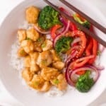A bowl of white rice topped with teriyaki chicken, broccoli, red bell peppers, red onions, sesame seeds, and chopped green onions, with chopsticks resting on the bowl.
