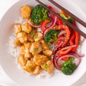 A bowl of white rice topped with teriyaki chicken, broccoli, red bell peppers, red onions, sesame seeds, and chopped green onions, with chopsticks resting on the bowl.